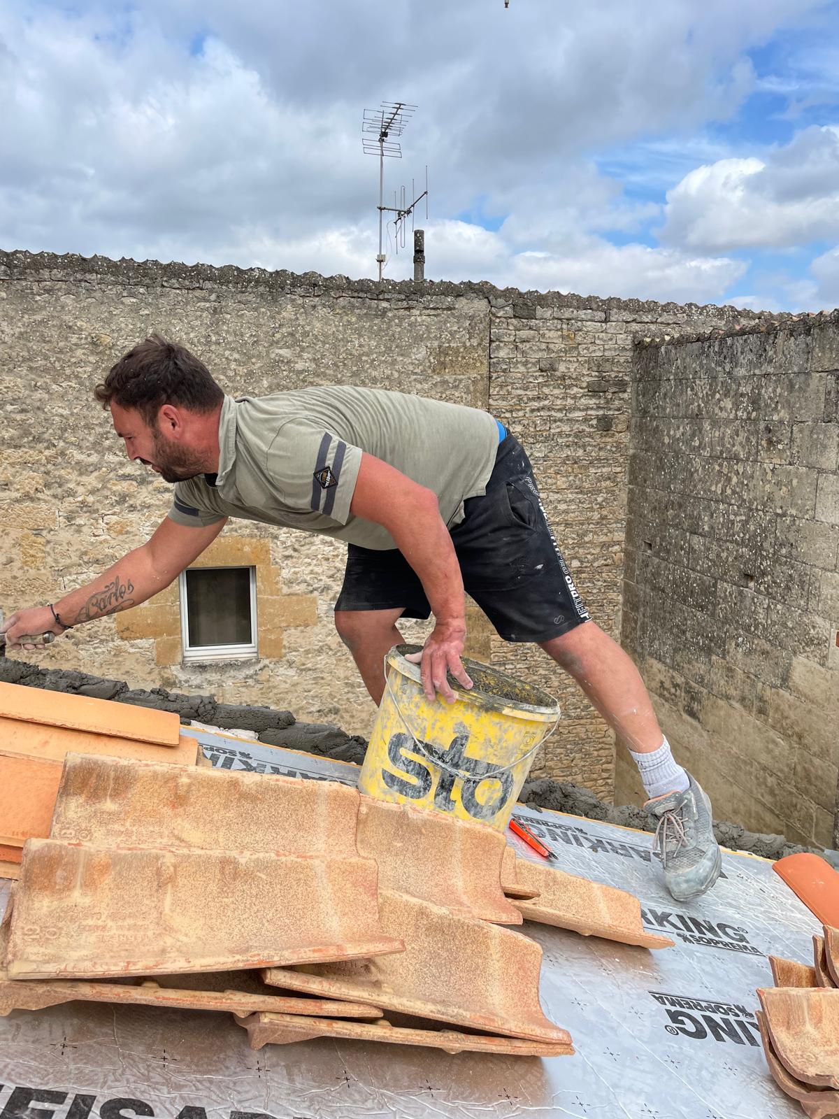Hermano, maçon, en train de poser des tuiles faîtières scellées au mortier sur une toiture, à Niort